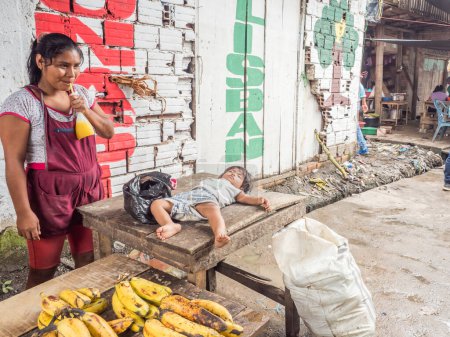 Foto de Belén, Iquitos, Perú - 27 de marzo de 2018: Mujer joven vendiendo plátanos en el mercado de Belén en Iquitos y el niño está durmiendo en la mesa de madera Beln. - Imagen libre de derechos