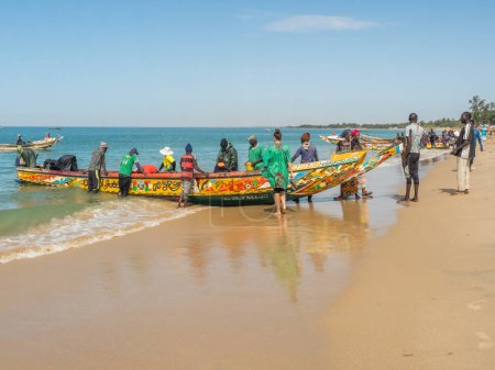Foto de Nianing, Senegal - 2 de enero de 2019: Pescadores recogiendo peces de madera de colores barco pescador de pie en la playa. África - Imagen libre de derechos