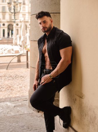 Atractivo atlético hombre con camisa abierta en el torso muscular desnudo, apoyado contra el pilar, en el centro de la ciudad europea, con camisa y pantalones negros