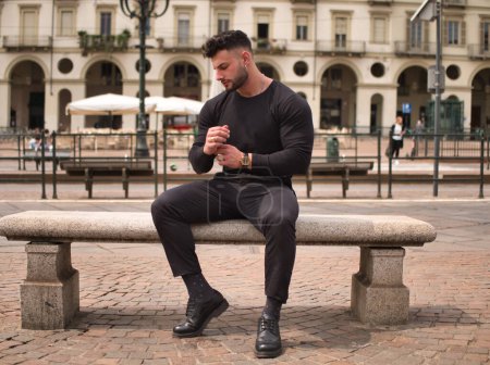 Atractivo atleta sentado en un banco de piedra en el centro de la ciudad, con camisa y pantalones negros