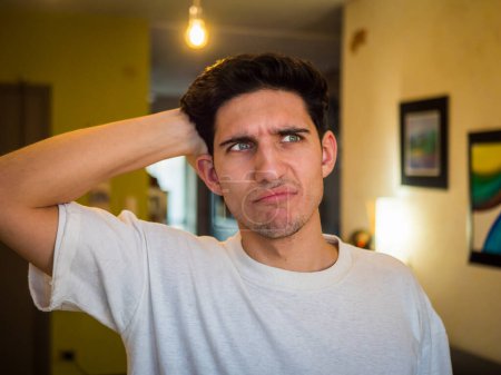 Photo for Confused or doubtful young man scratching his head and looking up. Indoors shot in a living room - Royalty Free Image