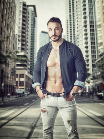 Foto de Handsome young man standing in the middle of a city street, looking at camera, wearing a shirt open on naked muscular torso - Imagen libre de derechos