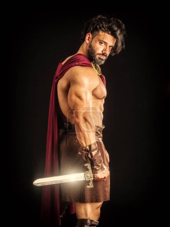 Photo for Handsome muscular man posing in roman or spartan gladiator costume with shield and sword, isolated on black background in studio - Royalty Free Image