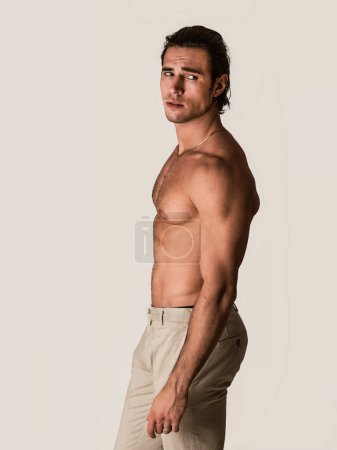 Photo for Three quarters, profile shot of handsome shirtless athletic young man in pants, looking at camera in studio shot - Royalty Free Image