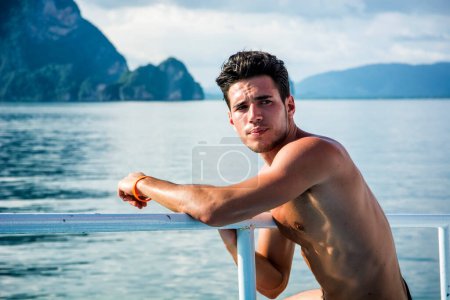 Photo for Portrait of young handsome bare-chested brunet man looking away against seascape on a boat or ship, leaning on handrail. - Royalty Free Image