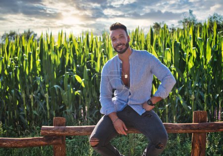 Photo for Handsome Hunky Young Man Outdoor in Countryside Sitting on Wood Fence, While Looking at Camera - Royalty Free Image