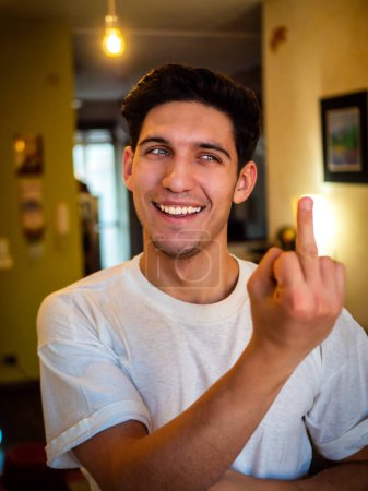 Photo for Young man doing screw you sign with middle finger looking away to a side with a smile, indoor at home - Royalty Free Image