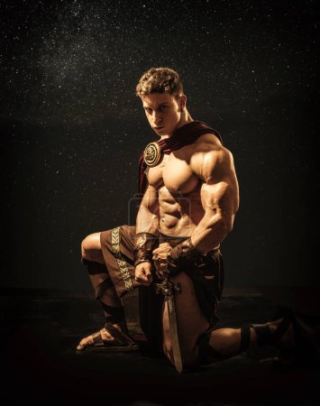Photo for Handsome muscular young man posing kneeling in roman or spartan gladiator costume with shield, on black background in studio - Royalty Free Image