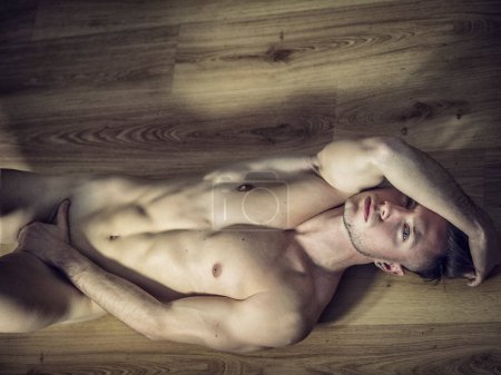 Guapo totalmente desnudo musculoso joven acostado en el suelo de madera en casa en actitud seductora, mirando a la cámara
