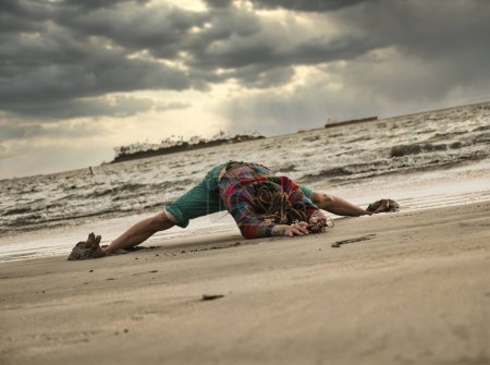 Téléchargez les photos : Pleine longueur anonyme gars avec dreadlocks faire large pattes avant courber sur le sable humide près de la mer ondulante dans la soirée nuageuse - en image libre de droit