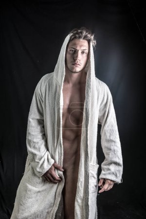 Téléchargez les photos : Beau jeune homme totalement nu portant un peignoir blanc, sur fond sombre en studio tourné - en image libre de droit