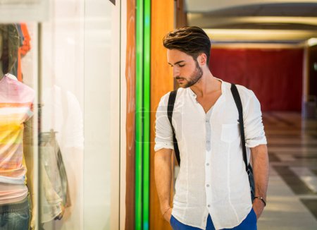 Foto de Hombre joven guapo con camisa blanca y mochila mirando los artículos de moda exhibidos en la boutique de la ventana de cristal en el lado de la calle. - Imagen libre de derechos