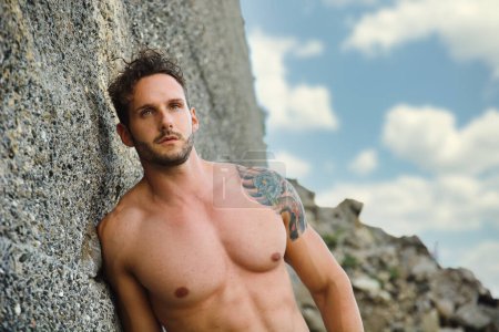 Photo for Attractive muscular shirtless athletic man standing on rock next to water by sea or ocean shore, looking at camera in a cloudy summer day - Royalty Free Image