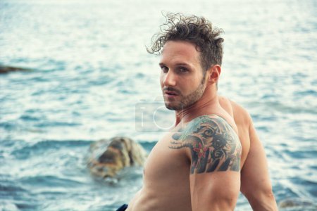 Photo for Attractive muscular shirtless athletic man sitting next to water by sea or ocean shore, looking at camera in a cloudy summer day - Royalty Free Image