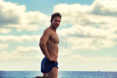 Photo for Attractive muscular shirtless athletic man standing next to water by sea or ocean shore, looking at camera in a cloudy summer day - Royalty Free Image