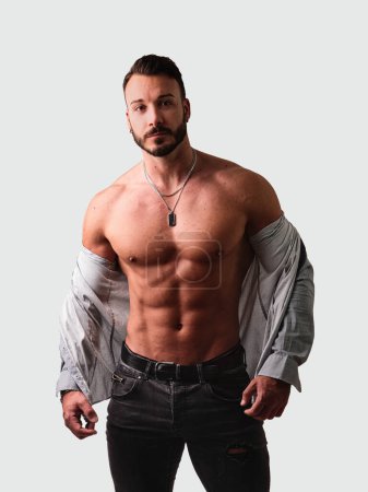 Handsome young muscular man shirtless wearing jeans, taking off white shirt on naked muscle torso, on light background in studio shot