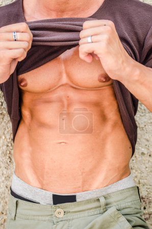 Foto de Sexy joven musculoso hombre levantando camisa para mostrar abdominales y pectorales, tiro al aire libre - Imagen libre de derechos