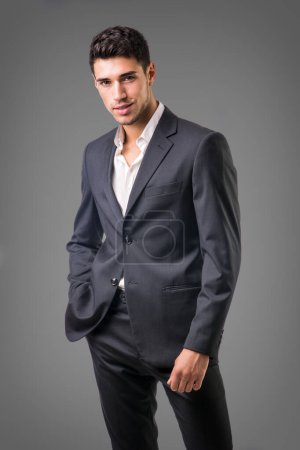 Jeune homme d'affaires posant en toute confiance devant la caméra, portant un costume d'affaires sans cravate, avec chemise ouverte sur le cou, sur fond sombre