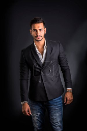 Photo for Young businessman confidently posing in front of camera, wearing business suit without neck-tie, with shirt open on neck, on dark background - Royalty Free Image