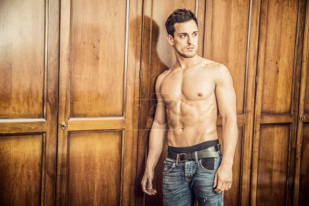 Téléchargez les photos : Sexy beau jeune homme debout torse nu dans sa chambre à coucher contre la porte de la penderie en bois - en image libre de droit