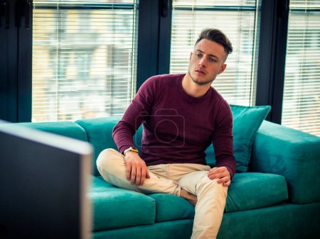 Photo for Young man sitting watching television changing the channel with the remote control - Royalty Free Image