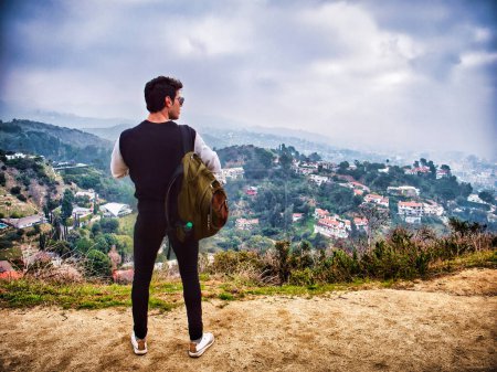 Téléchargez les photos : Stylish young man with backpack and photo camera, a photographer or a tourist, looking away over shoulder while standing against green hills over Los Angeles, California, with houses on foggy day - en image libre de droit