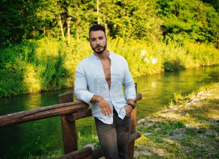 Téléchargez les photos : Beau jeune homme beau en plein air dans la campagne penché sur la clôture en bois, tout en regardant loin - en image libre de droit