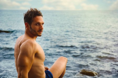 Photo for Attractive muscular shirtless athletic man sitting next to water by sea or ocean shore, looking at camera in a cloudy summer day - Royalty Free Image