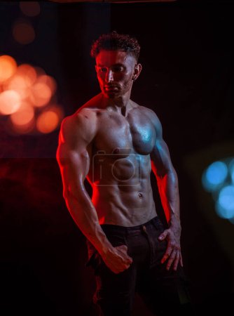 In einem Studio-Setting steht ein auffälliger männlicher Bodybuilder im Rampenlicht. Die Beleuchtung erzeugt eine dramatische Wirkung, die die Konturen seiner wohldefinierten Muskeln betont. Er posiert selbstbewusst