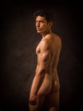Photo for Portrait of naked handsome young man with languishing look covering crotch with his hands. In a studio with a dark backdrop, an athletic, handsome young man stands completely nude looking at camera. - Royalty Free Image