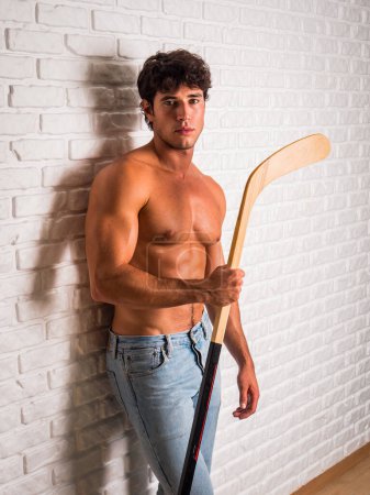 Foto de Joven guapo sin camisa con palo de hockey en las manos apoyado contra la pared de ladrillo blanco, mirando a la cámara, usando jeans - Imagen libre de derechos