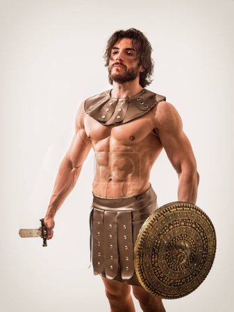 Photo for A handsome and well-built man strikes a pose while shirtless, dressed in a Roman or Spartan gladiator costume, set against a neutral grey backdrop. Young handsome muscular man posing shirtless in roman or spartan gladiator costume in studio. - Royalty Free Image