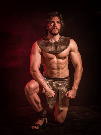 Photo for Young handsome muscular man posing shirtless in roman or spartan gladiator costume in studio. A handsome and well-built man strikes a pose while shirtless, dressed in a Roman or Spartan gladiator costume, set against a black backdrop. - Royalty Free Image