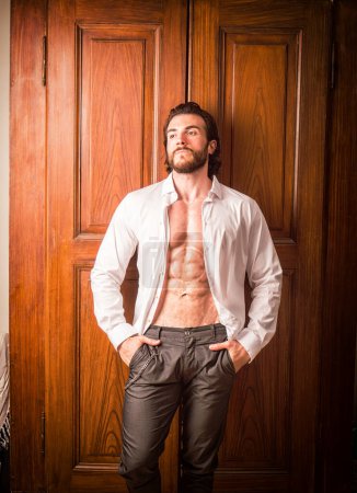 Téléchargez les photos : Sexy bel homme debout en chemise ouverte blanche avec un sourire devant les portes du placard en bois - en image libre de droit