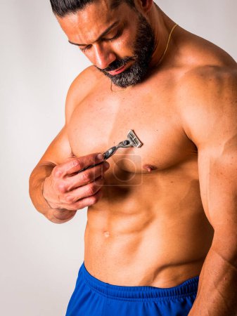 A shirtless man with a razor blade in his hand to cut his chest hair and remove it