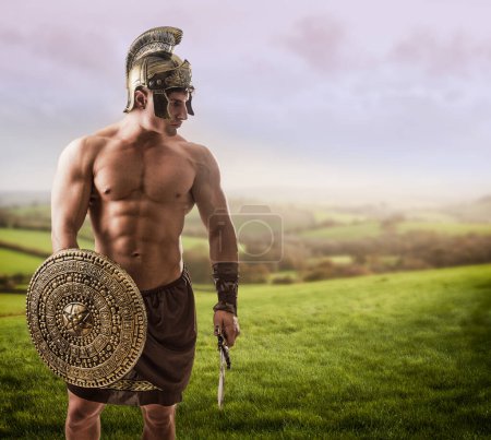 Photo for Young handsome muscular man posing in roman or spartan gladiator costume with shield and sword on field - Royalty Free Image