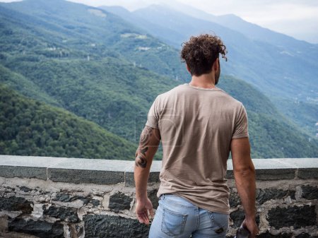 unrecognizable man seen from the back, standing by a stone wall with a scenic background, looking away at the view.