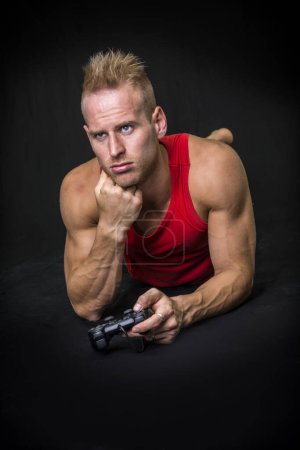 Téléchargez les photos : Un homme dans un débardeur rouge étendu sur le sol, ennuyé, avec un joystick de jeux vidéo à la main - en image libre de droit