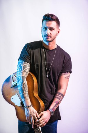 Photo for A young handsome man holding a guitar in his right hand in studio shot on neutral background. Photo of a man playing a guitar with passion and skill - Royalty Free Image