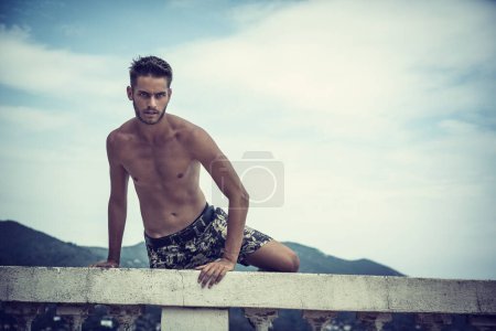 Photo for A shirtless man sitting on a ledge. Photo of a handsome man with captivating blue and green eyes sitting on a ledge - Royalty Free Image