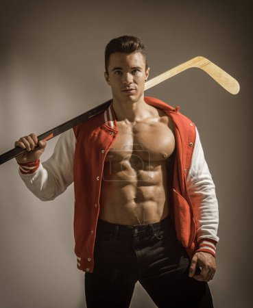 Foto de Un joven musculoso sosteniendo un palo de hockey y usando una chaqueta roja abierta en el torso muscular. Un hombre fuerte listo para jugar hockey - Imagen libre de derechos