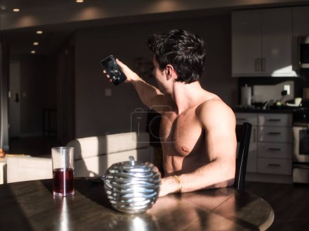 Foto de Un joven musculoso sin camisa sentado en una mesa tomando una foto de sí mismo. Foto de un hombre capturando un autorretrato en una mesa a la luz de la mañana - Imagen libre de derechos