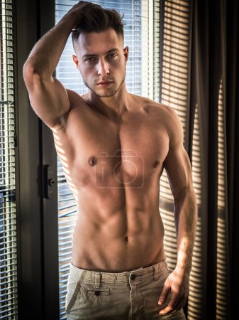 Portrait of sexy shirtless man covered with shadow stripes from window, looking at camera