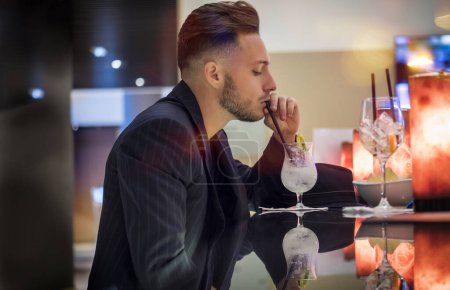 Photo for Handsome young man drinking cocktail at bar counter, wearing business suit and looking at camera - Royalty Free Image