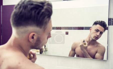 Téléchargez les photos : Bel homme jeune torse nu dans sa salle de bain maison, pulvérisation d’eau de cologne ou de parfum sur le cou - en image libre de droit