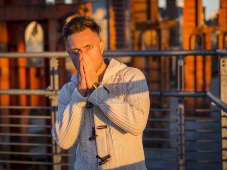 Man in white sweater covering mouth with hands and looking shocked at camera standing in sunlight on street 