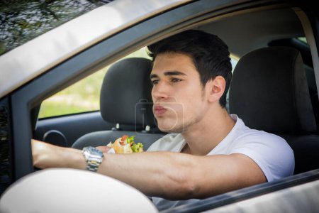 Téléchargez les photos : Jeune bel homme conduisant sa voiture tout en mangeant de la nourriture dans la circulation - en image libre de droit