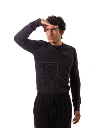Ein Mann in einem lässigen schwarzen langärmeligen Hemd und dunkler Hose steht mit der linken Hand über der Stirn, als würde er seine Augen vor Blendung schützen, während er aufmerksam auf etwas in der Ferne Unsichtbares blickt..