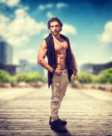 A shirtless man stands confidently on a brick road, the sunlight casting shadows across his muscular frame. The urban setting contrasts with his natural presence.