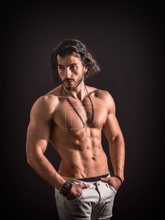 Un hombre con el pelo largo está de pie sin camisa, haciendo una pose para una foto. Su físico tonificado se destaca mientras mira con confianza a la cámara.
